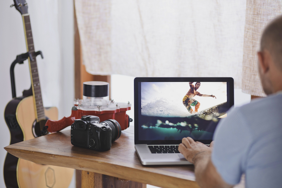 Cámara Canon en el escritorio al lado de la persona que edita imágenes de surf en una computadora portátil con una unidad de vivienda submarina en el fondo.